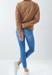 Béžová semišová bunda se zipy na rukávech, kombinovaná s modrými úzkými džíny a bílými teniskami. Zvýrazněné čisté linie a minimální doplňky.