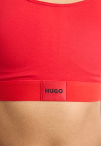 Reggiseno sportivo rosso con scollatura arrotondata e fascia elastica che presenta il logo "HUGO" in nero. Materiale liscio ed elastico, design aderente.