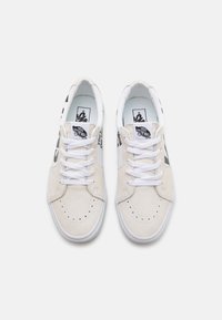 Beżowe zamszowe trampki z białymi sznurowadłami, okrągłym noskiem, perforowanymi detalami, czarnymi akcentami z logo i białą gumową podeszwą.