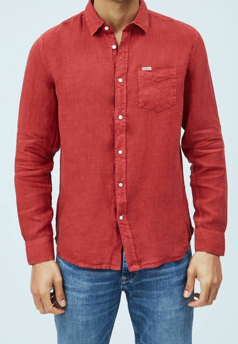 Homme portant une chemise rouge à manches longues avec des boutons et une poche poitrine, ainsi qu'un jean bleu, debout devant un fond clair uni.