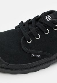 Svart kanvas sneakers med snøring, metallhull, gummisåle og "PALLADIUM" logo etikett på siden. Teksturert overmateriale.