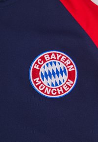 Izvezeni logo FC Bayern Münchena na tamnoplavoj pozadini, kružni dizajn sa crvenim obrubom, plavi i bijeli dijamantni uzorak, te bijelim tekstom.