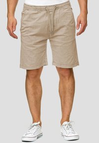 Shorts en coton beige avec une taille élastique et un cordon de serrage, dotés de poches latérales et d'une coupe décontractée. Portés avec des baskets blanches.