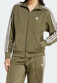 Olivově zelená sportovní bunda se zipem vpředu, bílými pruhy a bočními kapsami. Vyrobena z lesklé tkaniny, s vyšitým logem na hrudi.