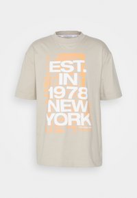 Beige katoenen T-shirt met korte mouwen en ronde hals. Heeft witte en oranje tekst: "EST. IN 1978 NEW YORK" in grafisch ontwerp.