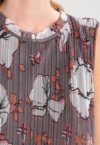 Hihaton harmaa pliseerattu paita, jossa on suuria valkoisia ja oransseja perhosiprinttejä sekä rypytetty, kerroksellinen kaulus.