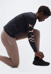 Άτομο γονατιστό δένοντας μαύρο παπούτσι για τρέξιμο, φορώντας καφέ κολάν Salomon και μαύρο μακρυμάνικο μπλουζάκι με ροζ και κίτρινα σχέδια στο μανίκι.