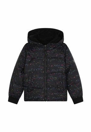 Joven chica de pie con las manos en los bolsillos, lleva una chaqueta acolchada negra, una sudadera con capucha rosa brillante, leggings negros y zapatillas blancas.