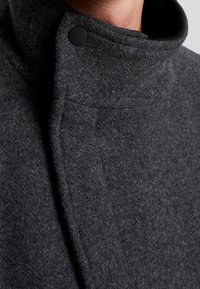 Cappotto di lana grigio con colletto alto, caratterizzato da una trama sottile e una chiusura a bottone nera vicino al collo. Design semplice e lineare.