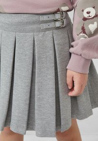 Kind trägt einen grauen Faltenrock mit zwei seitlichen Schnallen und einen rosa Pullover mit einem Teddybär-Design, die Hand entspannt an der Seite.