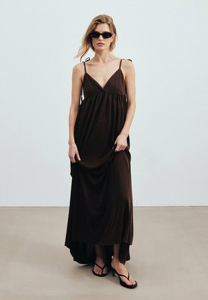 Woman wearing a long dark brown sleeveless dress, black sunglasses, and black heeled sandals standing indoors against a plain wall.