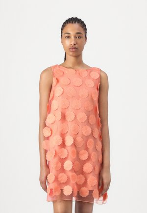 Woman with braided hair wearing sleeveless sheer coral dress decorated with large circular floral appliques, standing against a plain background.