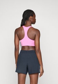 Femme avec des cheveux tressés, portant un soutien-gorge de sport rose vif et un short de sport noir, vue de dos sur un fond uni.