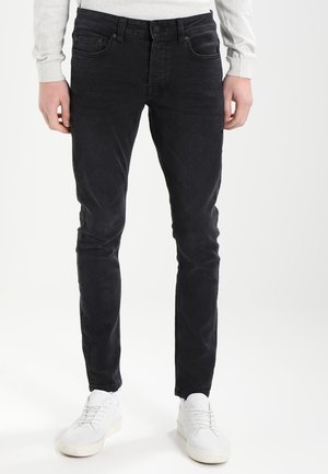 Muž v černých úzkých džínách, bílých teniskách a světle šedém tričku s dlouhým rukávem stojí na jednobarevném bílém pozadí.