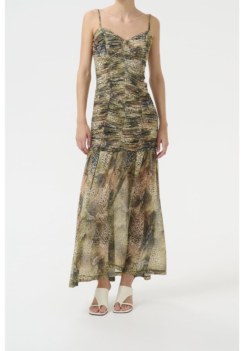 Femme portant une robe maxi sans manches à motif serpent avec de fines bretelles et des sandales blanches à talons ouverts, se tenant sur un fond uni.