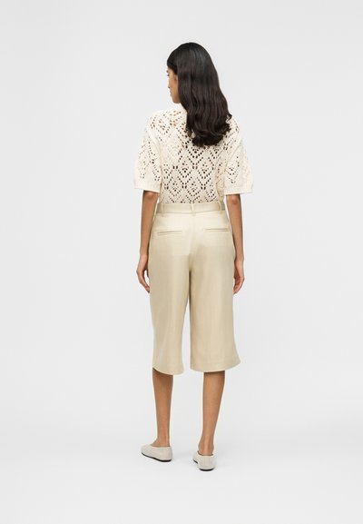 Femme aux cheveux longs et bruns portant un haut en crochet crème à manches courtes, un pantalon beige longueur genou et des chaussures plates beiges, debout dos à la caméra.