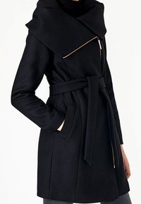 Cappotto in misto lana nero con ampio colletto pieghettato, chiusura a zip obliqua, due tasche frontali e cintura annodata in vita.