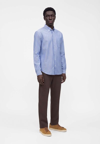 Homme debout portant une chemise bleu clair boutonnée, un pantalon marron foncé et des baskets en daim beige sur fond blanc.