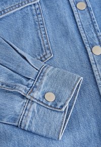 Tissu en denim bleu clair avec boutons-pression métalliques, montrant un poignet et une partie de poche avec des coutures orange visibles.