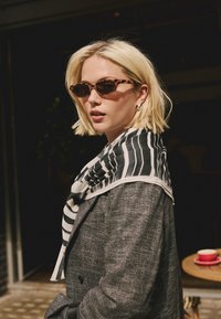 Braune Schildpatt-Sonnenbrille, schwarz-weiß gestreifter Schal, grauer strukturierter Mantel, kurze blonde Haare, stehend vor dunklem Hintergrund.