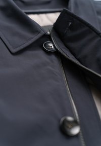 Nahaufnahme einer dunklen Jacke mit Kragen, teilweise offenem Reißverschluss und zwei großen schwarzen Knöpfen auf glattem Stoff.