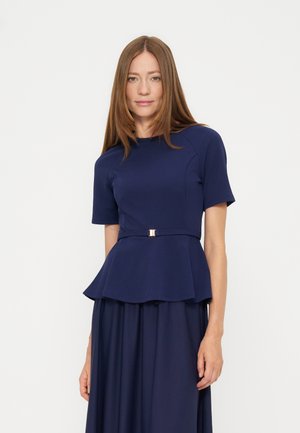 Frau mit langen glatten Haaren, die ein marineblaues kurzärmeliges Peplum-Oberteil und einen passenden Rock trägt, vor weißem Hintergrund stehend.