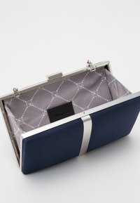 Navy blue kopertówka z metalową ramką. Wnętrze wykończone szarym materiałem z wzorzystym deseniem. Zawiera detal z logo na materiałowej metce.