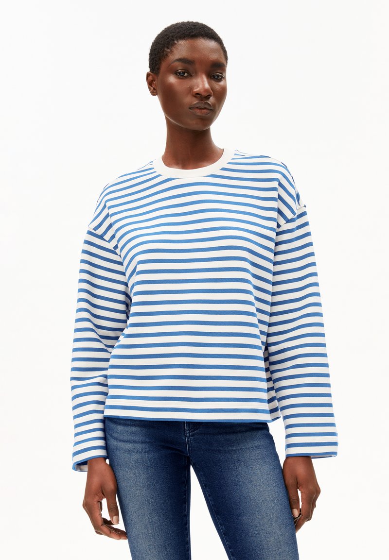 Donna che indossa una camicia a maniche lunghe a righe orizzontali blu e bianche e jeans blu, in piedi contro uno sfondo bianco semplice.