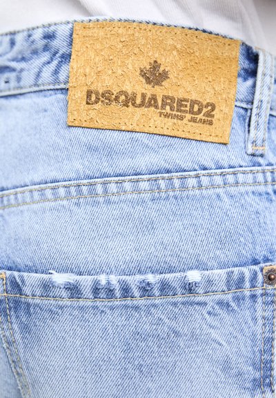 Un bolsillo de unos vaqueros de denim azul claro con un parche de cuero beige etiquetado "Jeans de los gemelos DSQUARED2" y un pequeño detalle deshilachado.