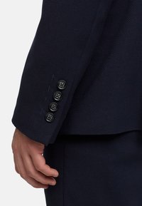 Marineblauwe gestructureerde blazer manchet met vier zwarte knopen met een bloemmotief, toont een gestructureerde mouw en subtiele stofweefdetails.