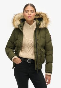 Groene pufferjack met een met bont gevoerde capuchon, ritssluiting, zijzakken, gedragen over een crèmekleurige kabelgebreide trui en zwarte broek.