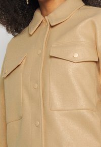 Beżowa koszula zapinana na guziki o miękkiej teksturze, z kołnierzykiem i dwoma kieszeniami na piersi z dopasowanymi guzikami.