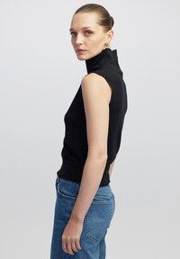 Top nero senza maniche a collo alto realizzato in tessuto a maglia, caratterizzato da una vestibilità aderente, superficie testurizzata e orlo sfilacciato. Abbinato a un paio di jeans blu a vita alta.