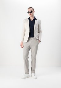 Lys beige blazer med en enkelt knapp, marineblå teksturert polo skjorte, lys grå bukser og hvite joggesko. Runde svarte solbriller fullfører antrekket.