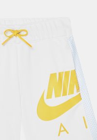 Bílé sportovní šortky s velkým žlutým logem Nike, hladkým povrchem, elastickým pasem se žlutou šňůrkou a síťovými bočními panely.