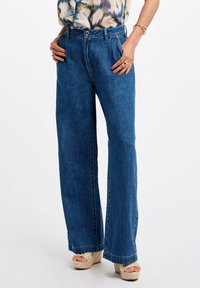 Bredde ben blå denim jeans med høy midje, med en klassisk fem-lomme design og subtil tekstur. Kombinert med kilehælsandaler.