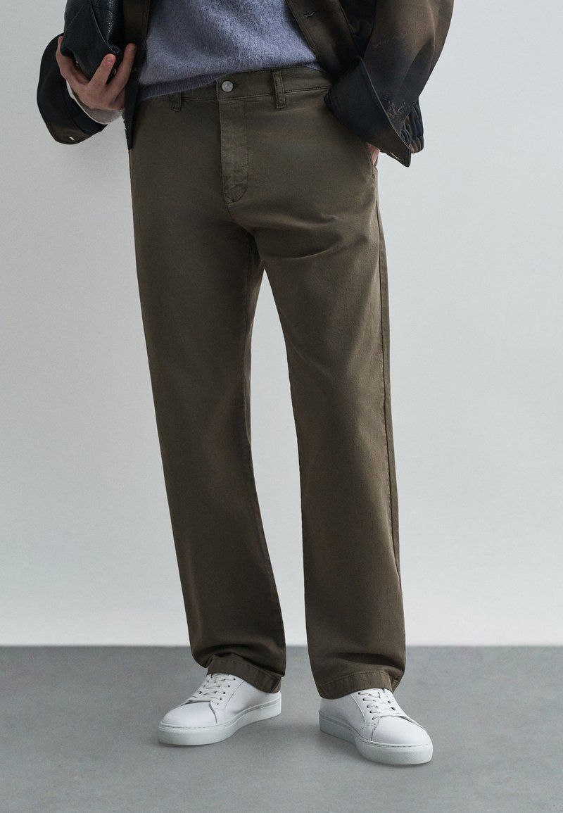 Καφέ βαμβακερές παντελόνες με χαλαρή εφαρμογή, σχέδιο πέντε τσεπών και ίσια πόδια, συνδυασμένες με λευκά sneakers για μια κομψή casual εμφάνιση.