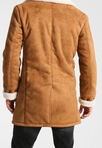 Brun shearlingjacka med vit fuskpälskrage och manschetter, med en slät yta och en strukturerad design, som sträcker sig till midjan.