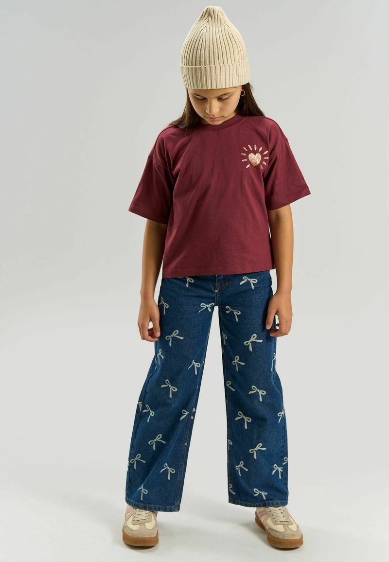 Enfant portant un bonnet beige en tricot, un t-shirt bordeaux avec un motif cœur pailleté, un jean large avec des motifs de nœuds blancs, et des baskets beiges, regardant vers le bas.
