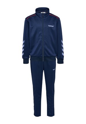 Dos niños con chándales deportivos azul marino están de pie uno frente al otro, uno con los brazos cruzados, usando zapatillas blancas y rosas respectivamente.
