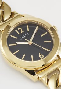 Montre en or avec un cadran noir et des index horaires en or. Dotée d'un bracelet en métal épais et d'une lunette ronde lisse. Nom de la marque "GUESS" affiché.
