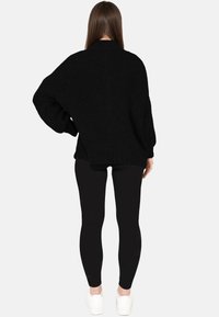 Kobieta o długich brązowych włosach ubrana w luźny czarny sweter, czarne legginsy i białe trampki, stoi tyłem z jedną ręką na biodrze.