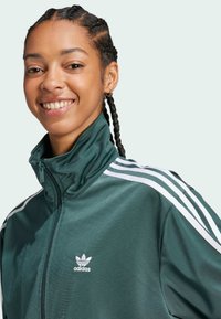Adidas track jacket in dark green, featuring white stripes on shoulders, high collar, and zip closure. Smooth fabric with logo detail.