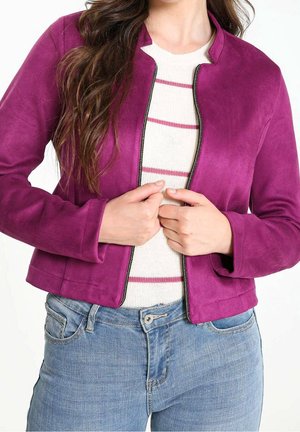 Kobieta w fioletowej kurtce na zamek błyskawiczny, na białym swetrze z różowymi paskami, w połączeniu z niebieskimi dżinsami, trzymająca boki kurtki.
