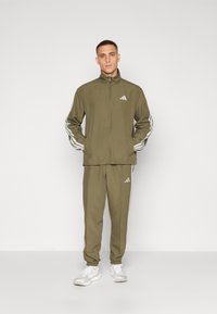 Tuta verde oliva realizzata in tessuto leggero, dotata di giacca con zip e righe bianche sulle maniche e pantaloni affusolati abbinati con dettagli.