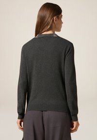 Grauer Pullover mit langen Ärmeln, strukturiertem Kragen und geripptem Saum, der eine glatte Stoffoberfläche aufweist. Getragen mit dunkelgrauen Hosen.