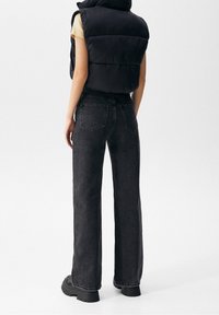 Osoba v černé zateplené vestě, béžovém tričku s krátkým rukávem, černých vysokých širokých džínách a robustních černých botách, stojící na bílém pozadí.