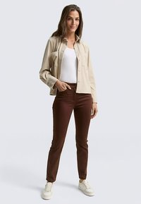 Béžová manšestrová košeľa na gombíky cez biele tielko, v kombinácii s hnedými džínsami a bielymi teniskami. Model stojí na neutrálnom pozadí.