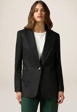 Schwarzer taillierter Blazer mit Spitzenrevers, einem Einzelknopfverschluss, zwei Vorderaschen und einer glatten Textur. Über einem weißen Oberteil und grünen Hosen getragen.