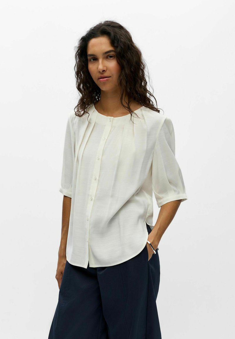 Femme aux cheveux bouclés foncés portant un chemisier blanc boutonné avec des manches trois-quarts et un pantalon ample foncé, debout avec une main dans la poche.